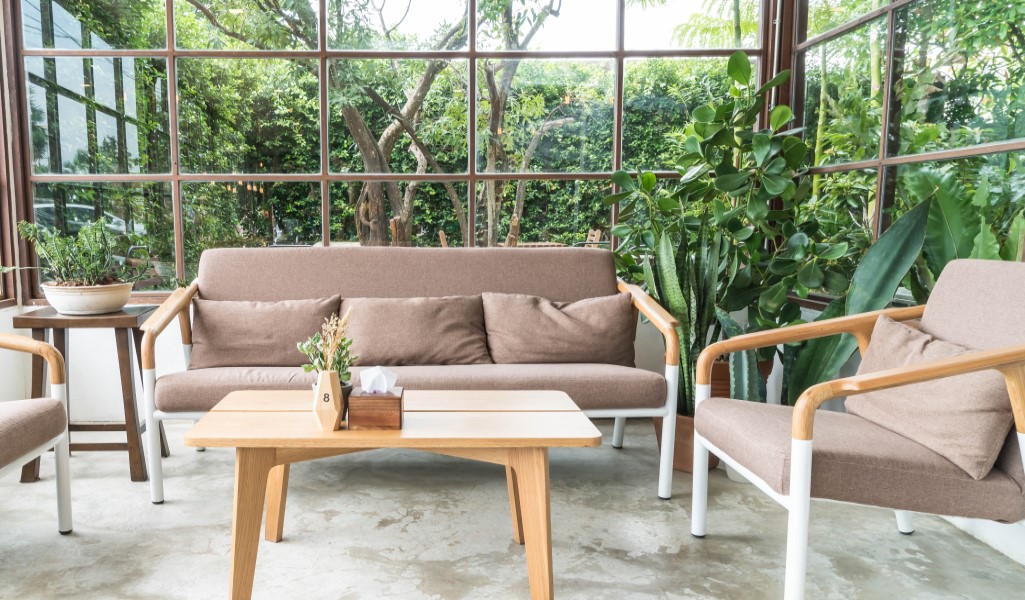 Porche cerrado con vidrio y vista al jardín, amueblado con sofás y sillones marrones, mesa de centro de madera y plantas decorativas.